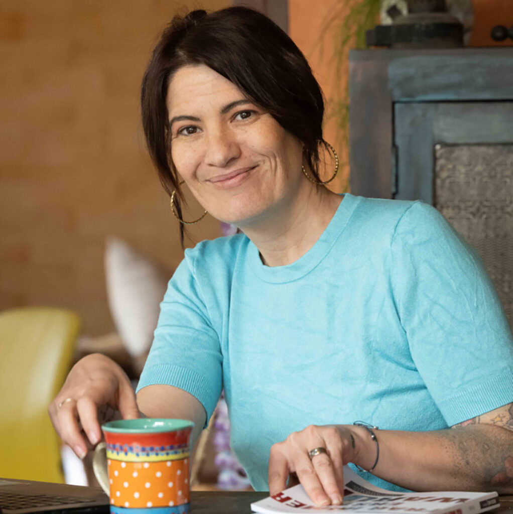 Vivien Schulter lächelnd am Schreibtisch mit einer Tasse, in einem hellen, einladenden Raum.