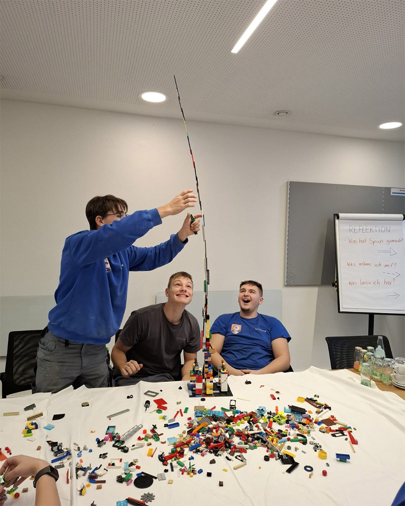 Mehrere Personen sitzen an einem Tisch voller LEGO Steine, eine Person baut einen sehr hohen LEGO-Turm, während die anderen lachen.