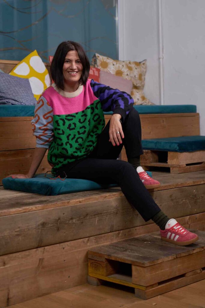Vivien Schulter sitzt entspannt auf Holzstufen, trägt einen bunten Pullover und lächelt in die Kamera.
