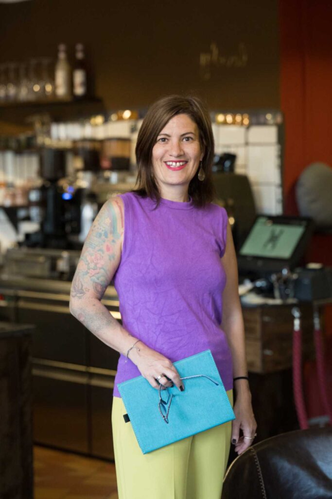 Vivien Schulter steht lächelnd in einem Innenraum und hält eine türkise Mappe und eine Brille in der Hand.