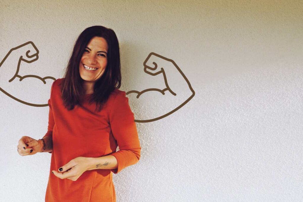 Vivien Schulter steht lächelnd vor einer hellen Wand, neben ihr sind zwei gezeichnete muskulöse Arme als Illustration zu sehen.